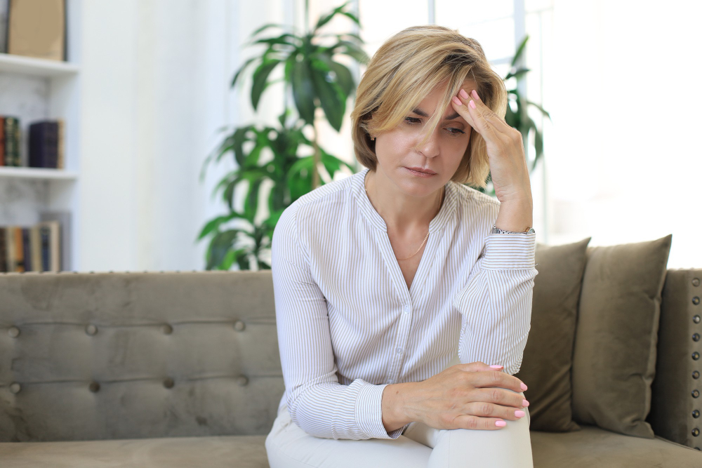 Middle-aged woman looking tired and stressed while sitting on a couch, representing perimenopause symptoms and women’s HRT in Parker CO