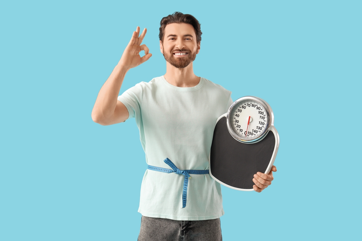 Smiling man holding a weight scale and giving an okay sign representing TRT and weight loss results in men with low testosterone